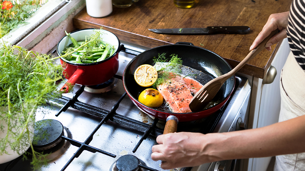 A woman cooking a salmon filet with lemon, fresh herbs, and green beans on the stove as part of her fertility diet. 