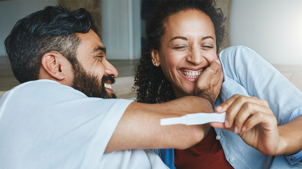 A man and woman smile joyfully as they look at a pregnancy test, potentially realizing they debunked common fertility myths.