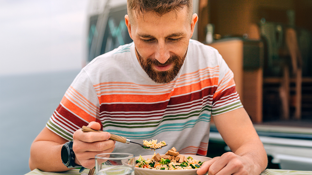 A bearded man in a striped t-shirt eats a veggie grain bowl with tofu and may be wondering about soy side effects in males.